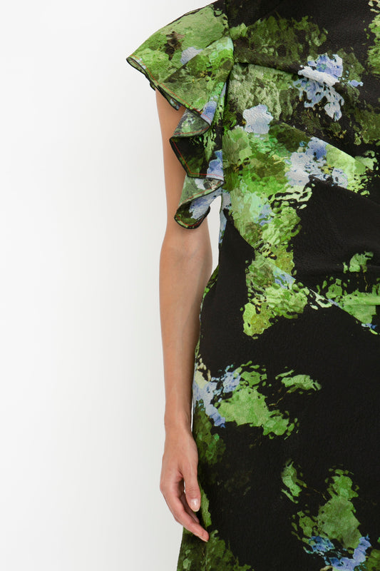 Close-up of a person wearing a green, black, and blue patterned Twist Shoulder Dress In Black Frost by Victoria Beckham with ruffled details on one shoulder and an asymmetric hemline, standing against a plain white background.