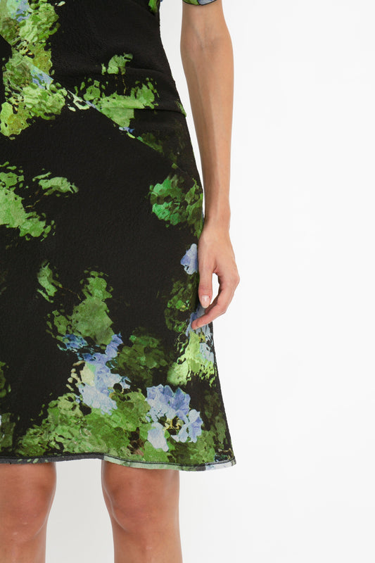 A close-up of a person wearing the Victoria Beckham Twist Shoulder Dress In Black Frost with a green and blue floral pattern, featuring draped pleat details. The person’s arm and part of their hand are visible.