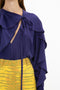 A person wears the Tie Detail Ruffle Blouse In Ultraviolet by Victoria Beckham, paired with high-waisted yellow patterned pants. The background is plain white.