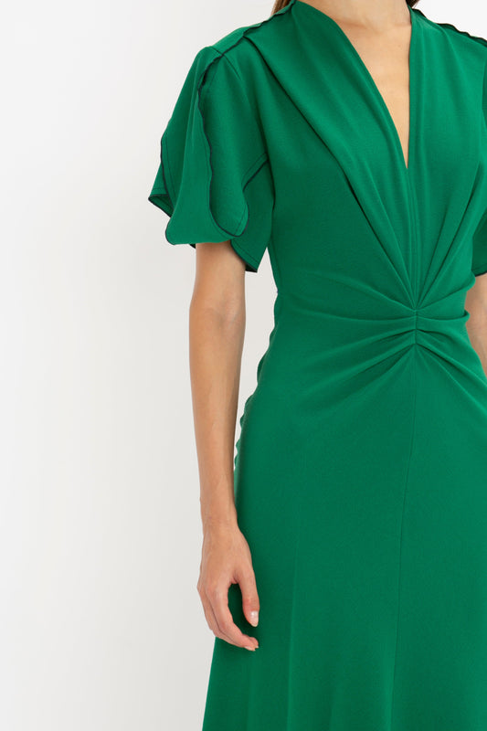 Close-up of a woman in a Victoria Beckham Gathered V-Neck Midi Dress in Emerald featuring flutter sleeves and a twisted detail at the chest, against a white background.