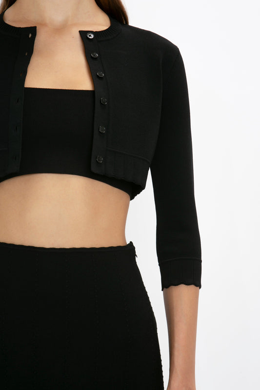 A woman is wearing the Victoria Beckham VB Body Cropped Cardigan In Black with feminine detailing over a black tube top, paired with a black high-waisted skirt that highlights her form-fitting silhouette.