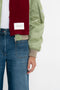 A person wearing a green jacket, an Exclusive Logo Patch Scarf In Burgundy by Victoria Beckham, and blue jeans stands against a plain background.