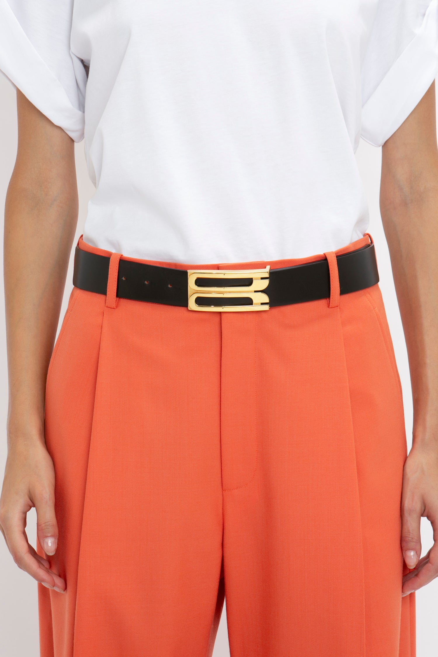 A person wearing orange pants with a Victoria Beckham Jumbo Frame Belt In Black Leather featuring gold hardware and a white t-shirt.