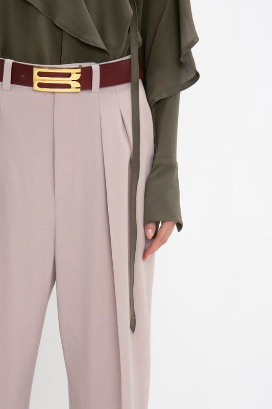 A person wearing the Victoria Beckham Double Pleat Trouser In Rose Quartz, a dark green top with long sleeves, and a maroon belt with a gold buckle showcases a sophisticated directional silhouette.