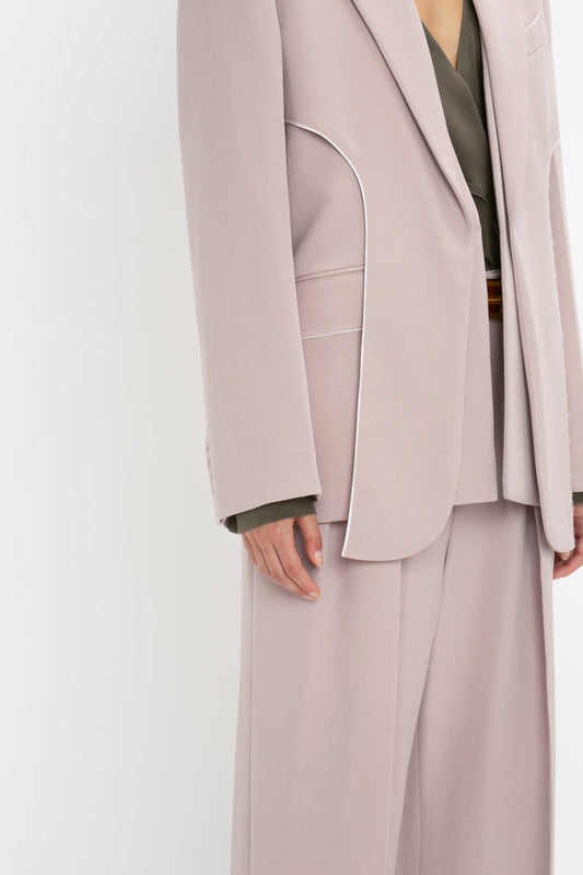 A person is shown wearing a beige blazer with white piping and matching pants, paired with a dark green top. The Victoria Beckham Double Panel Front Jacket In Rose Quartz adds an air of sophistication to the ensemble. The image focuses on the torso and upper legs without revealing the person's face.