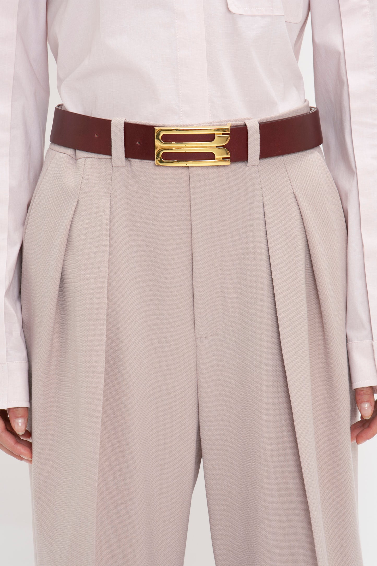 Close-up of a person wearing beige high-waisted pleated trousers, a white shirt, and a Jumbo Frame Belt In Burgundy Leather by Victoria Beckham with a gold rectangular buckle. The look is completed with contemporary Burgundy shoes made of calf leather.