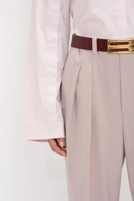 A person wearing a Button Detail Cropped Shirt In Rose Quartz from Victoria Beckham with high-waisted beige trousers and a red belt featuring a gold buckle.