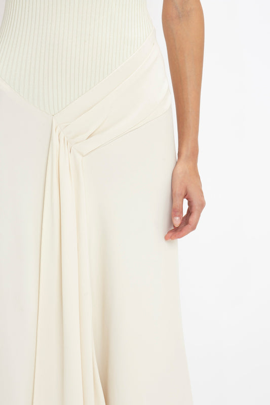 A person in a Victoria Beckham Sleeveless Tie Detail Dress In Cream stands with their arm resting at their side. The garment features a pleated draped detail at the waist and an elegant asymmetric hemline.