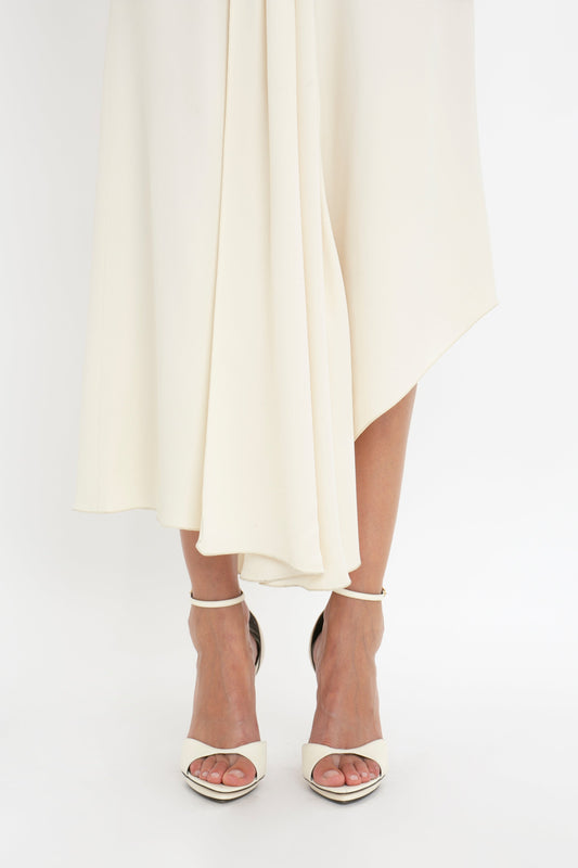 A close-up of a person wearing the Victoria Beckham Sleeveless Tie Detail Dress In Cream with an asymmetric hemline and white high-heeled sandals. The focus is on the lower part of the dress and feet.