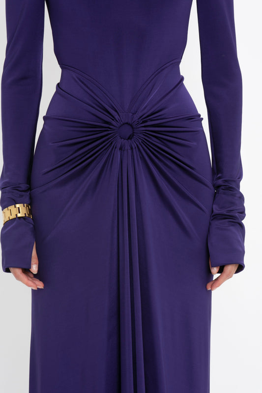 Close-up of a person wearing the Victoria Beckham Long Sleeve Gathered Midi Dress In Ultraviolet with intricate, body-sculpting stretch fabric detail at the waist, forming a central knot. The person is also wearing a chunky gold bracelet on the left wrist.