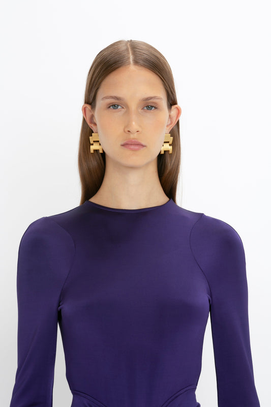 A person with long brown hair is wearing a purple Long Sleeve Gathered Midi Dress In Ultraviolet by Victoria Beckham and large geometric-shaped earrings. The background is plain white.