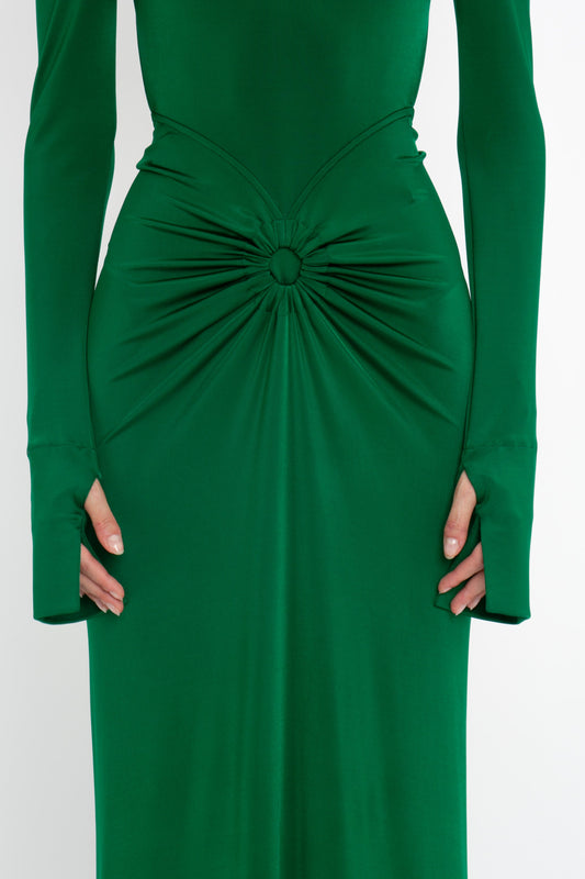 A close-up of a green dress with long sleeves, featuring a gathered and knotted detail at the waist. The hands of the person wearing the Circle Detail Open Back Gown In Emerald by Victoria Beckham are partially visible.