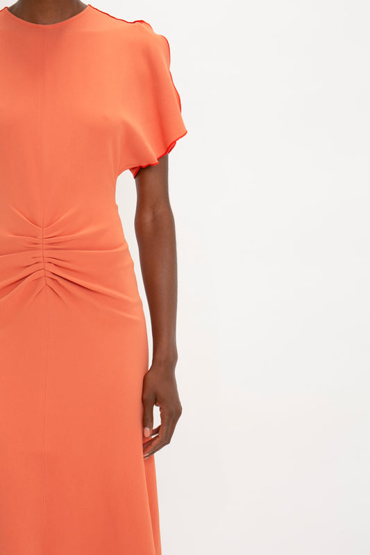A woman in a Victoria Beckham Gathered Waist Midi Dress In Papaya with a ruffled sleeve and centered twist detail, shown from the shoulders down to mid-thigh.