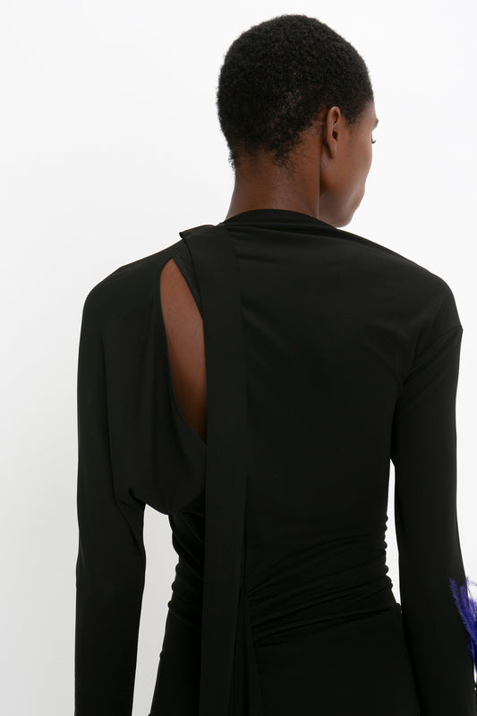 Rear view of a woman with short hair wearing a Victoria Beckham black Slash-Neck Ruched Midi Dress, with an asymmetric cut-out, standing against a white background.