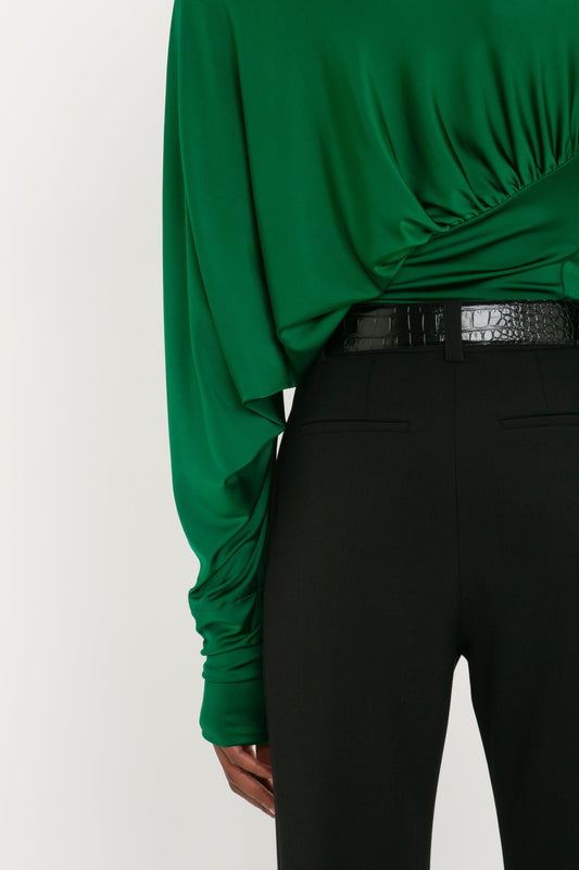 A person wearing a green long-sleeved top and Cropped Kick Trouser In Black from Victoria Beckham with a black belt. The image focuses on the lower back and waist area.