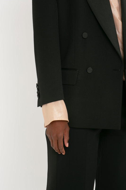 Close-up of a person wearing a Victoria Beckham Satin Lapel Tuxedo Jacket in Black over a light-colored shiny shirt. Only the lower part of the jacket, adorned with satin-covered buttons, and the person's hand are visible.