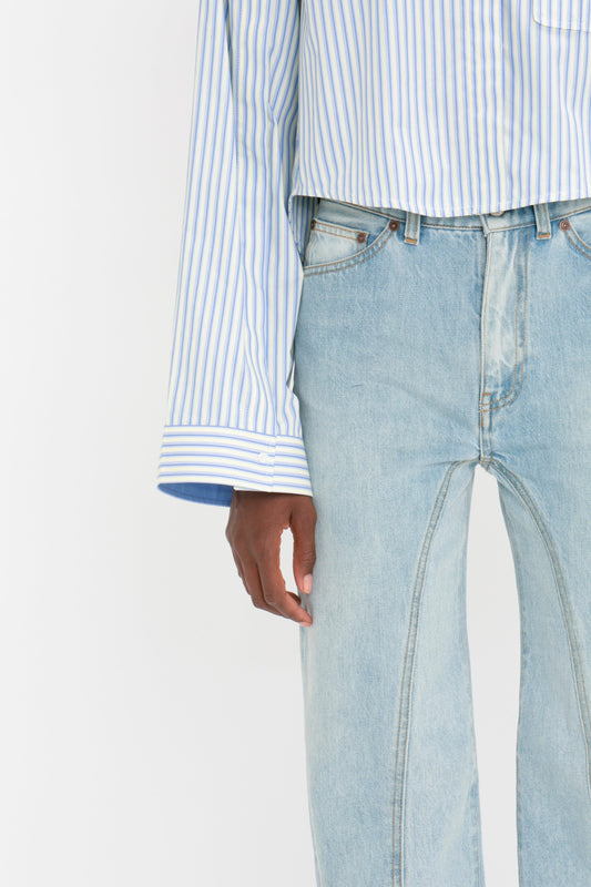 A person wearing a Victoria Beckham Button Detail Cropped Shirt In Chamomile Blue Stripe, paired with light blue jeans, exudes a chic and modern vibe. One arm hangs casually by their side, showcasing the outfit's elegant and feminine silhouette.