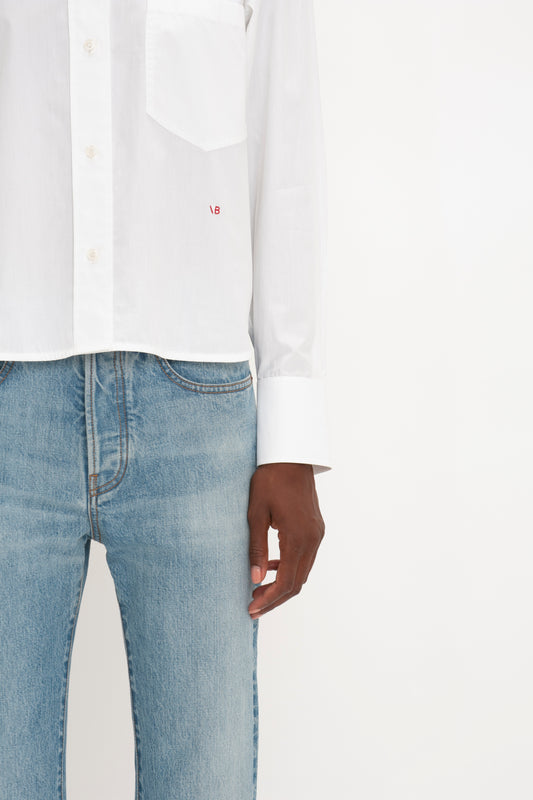 A person wearing a white Cropped Long Sleeve Shirt In White and blue jeans stands against a plain white background, showcasing Victoria Beckham's relaxed tailoring. Only the lower half of the shirt and the upper half of the jeans are visible.