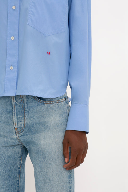 Close-up of a person wearing a light blue, relaxed fit Cropped Long Sleeve Shirt In Oxford Blue with a small red "VB" Victoria Beckham monogram on the chest pocket, made of organic cotton, paired with light blue jeans. The person's left hand is visible at their side.