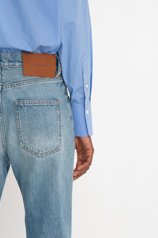 Close-up of a person wearing light blue, relaxed fit jeans with a leather label and a Victoria Beckham Cropped Long Sleeve Shirt In Oxford Blue with buttoned cuffs.