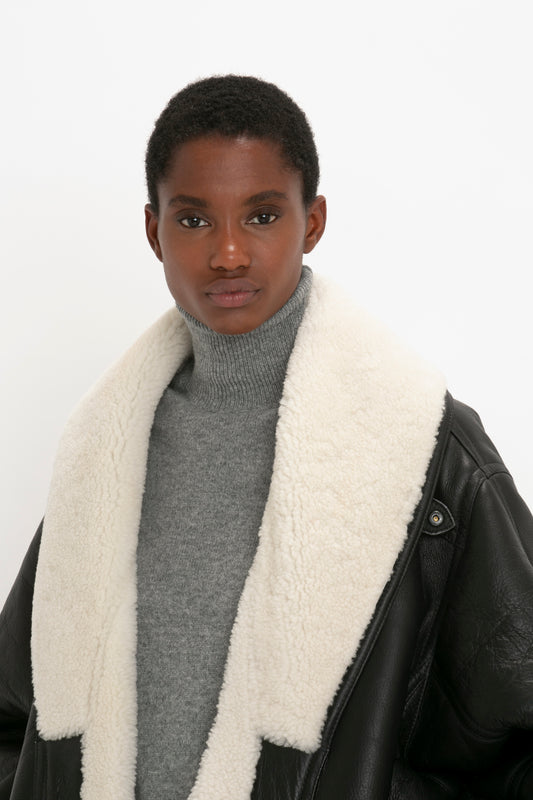 Person with short hair wearing a grey turtleneck sweater and an oversized silhouette black coat with a white shearling collar (Shearling Coat In Monochrome by Victoria Beckham), against a plain white background.
