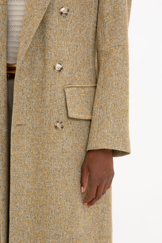 A close-up of a person’s hand resting by their side, wearing the Victoria Beckham Exclusive Waisted Tailored Coat In Flax with gray buttons over a textured beige shirt.