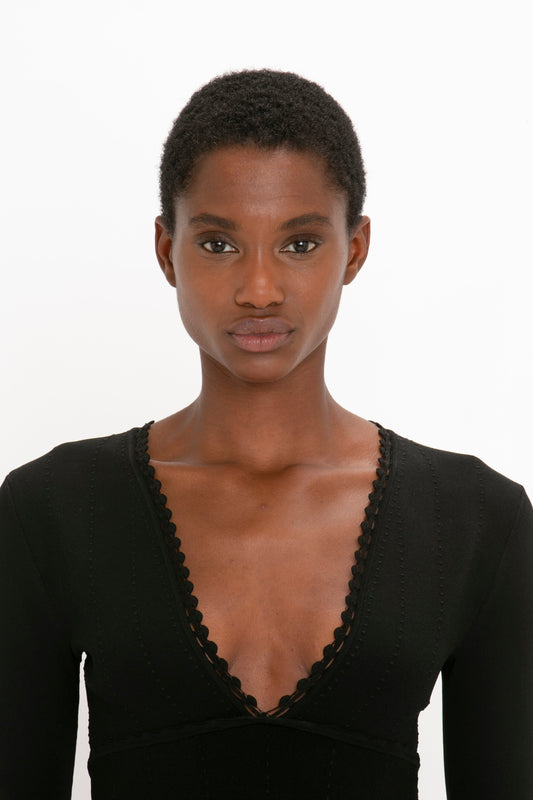 A person with short hair is standing against a plain white background, wearing the Victoria Beckham VB Body Long Sleeve V Neck Dress In Black.