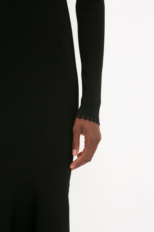 A close-up view of a person wearing a VB Body Long Sleeve V Neck Dress In Black by Victoria Beckham. Only the arm and hand are visible, with a scalloped edge detail on the cuff. The background is plain and white.