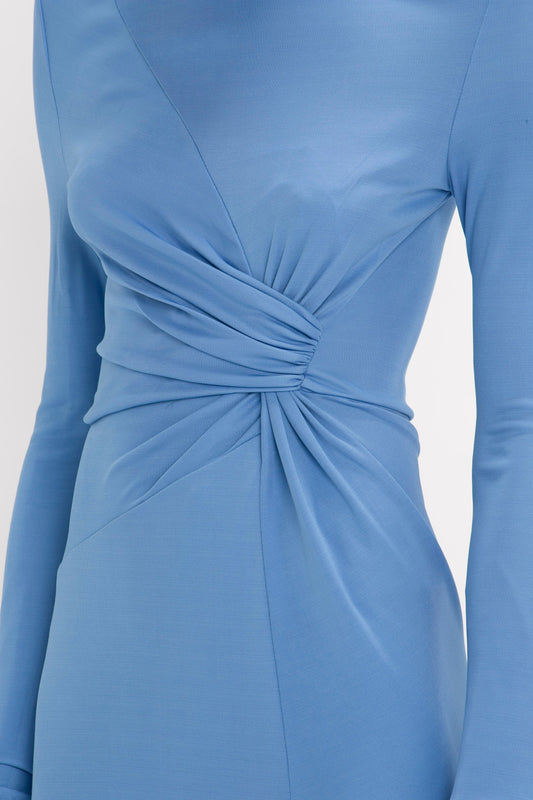 Close-up of a Victoria Beckham High Neck Asymmetric Draped Dress in Oxford Blue with long sleeves and a twisted fabric detail at the waist on a light grey background.