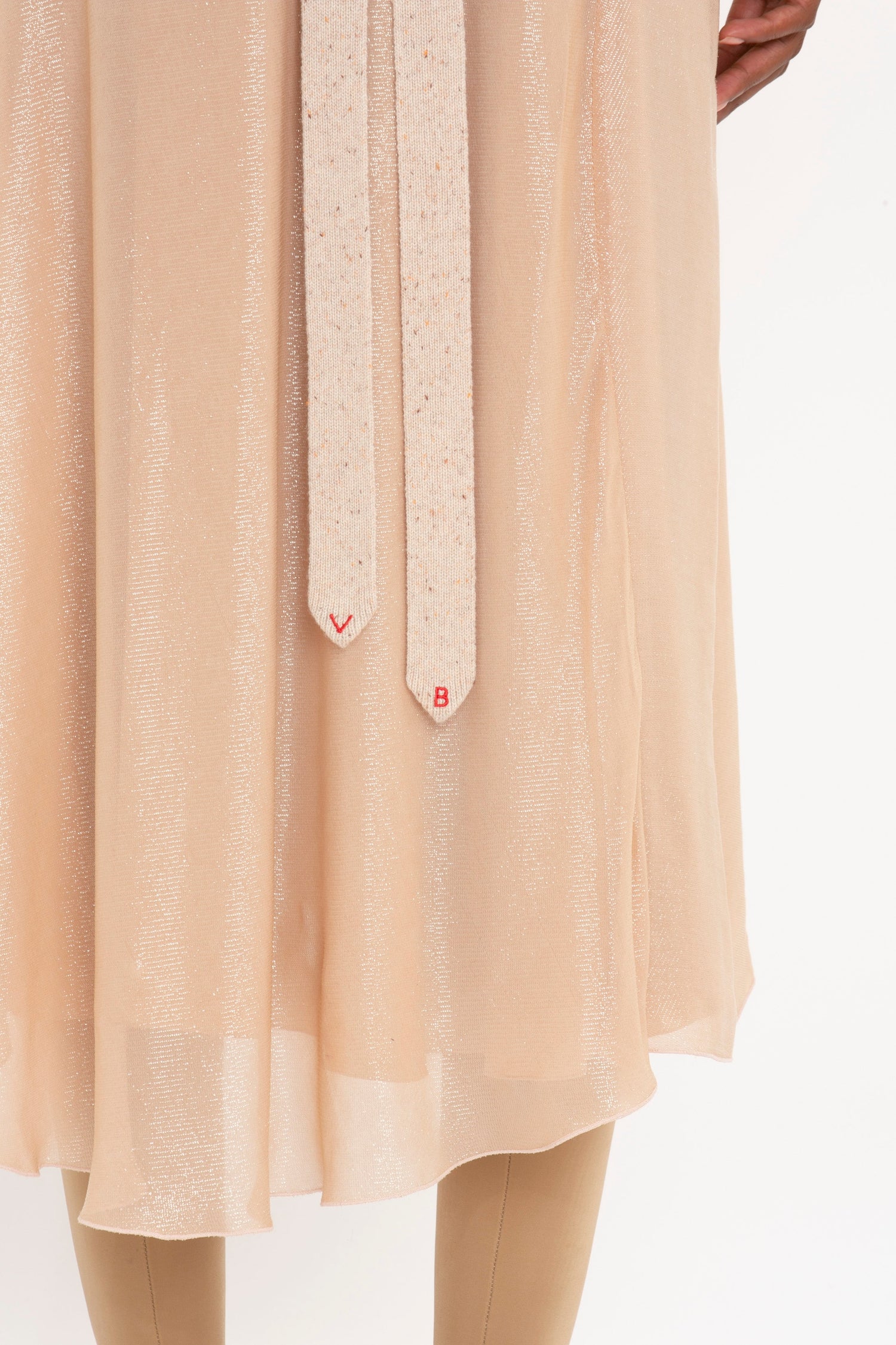 Close-up of a person's lower body in a beige outfit featuring the Victoria Beckham Wrap Front Cardigan In Nougat with a sheer, shimmery layer and beige pants. Two beige fabric strips with red stitching hang down, offering versatile styling options.