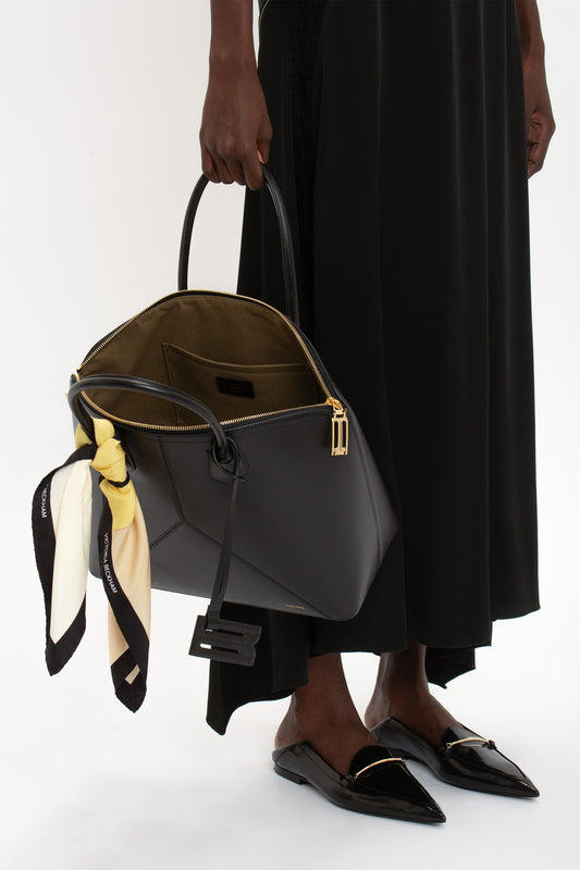 A person in a black dress holding a Victoria Beckham Victoria Bag In Black Leather with a yellow scarf tied to it, and wearing black pointed-toe shoes.