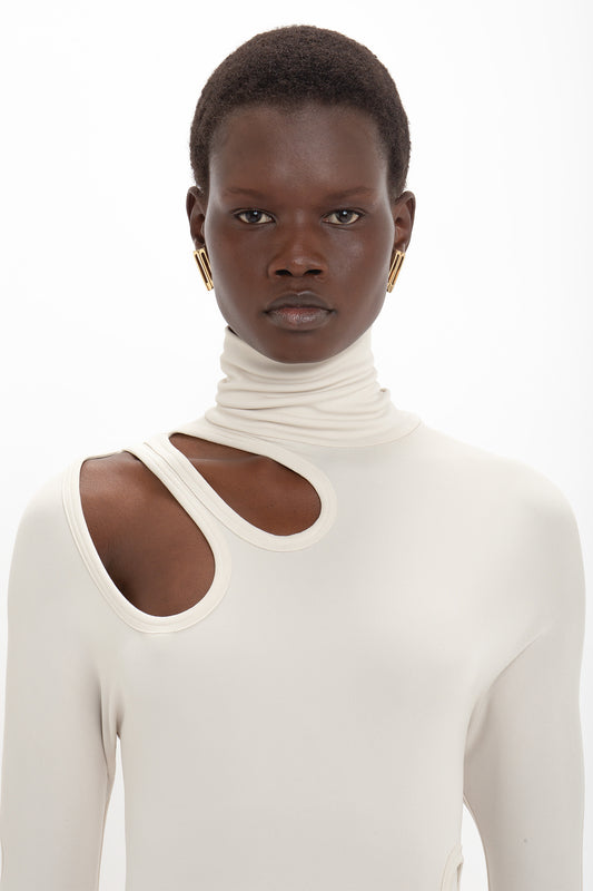 Person with short hair wearing a Victoria Beckham Long Sleeve Cut-Out Jersey Midi Dress In Bone, posing against a white background. They have gold hoop earrings.