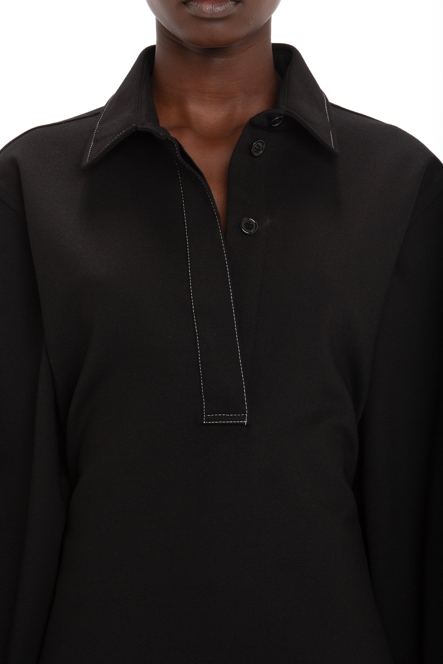 Close-up of a person wearing the Victoria Beckham Waistcoat Detail Ponti Top In Black with white stitching and a partially buttoned placket, showcasing a distinctive personality. The background is plain white.