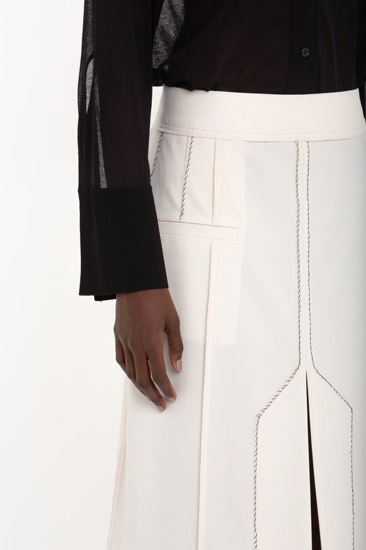A person wearing a black long-sleeve shirt with sheer details and a white skirt featuring vertical slits and contrast stitching, accessorized with the Pocket Detail Shirt In Black by Victoria Beckham for added flair.