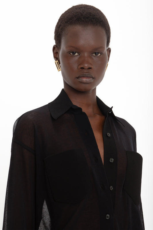 A person wearing a black, semi-sheer Pocket Detail Shirt In Black by Victoria Beckham with a collared neckline and gold earrings stands against a plain white background.