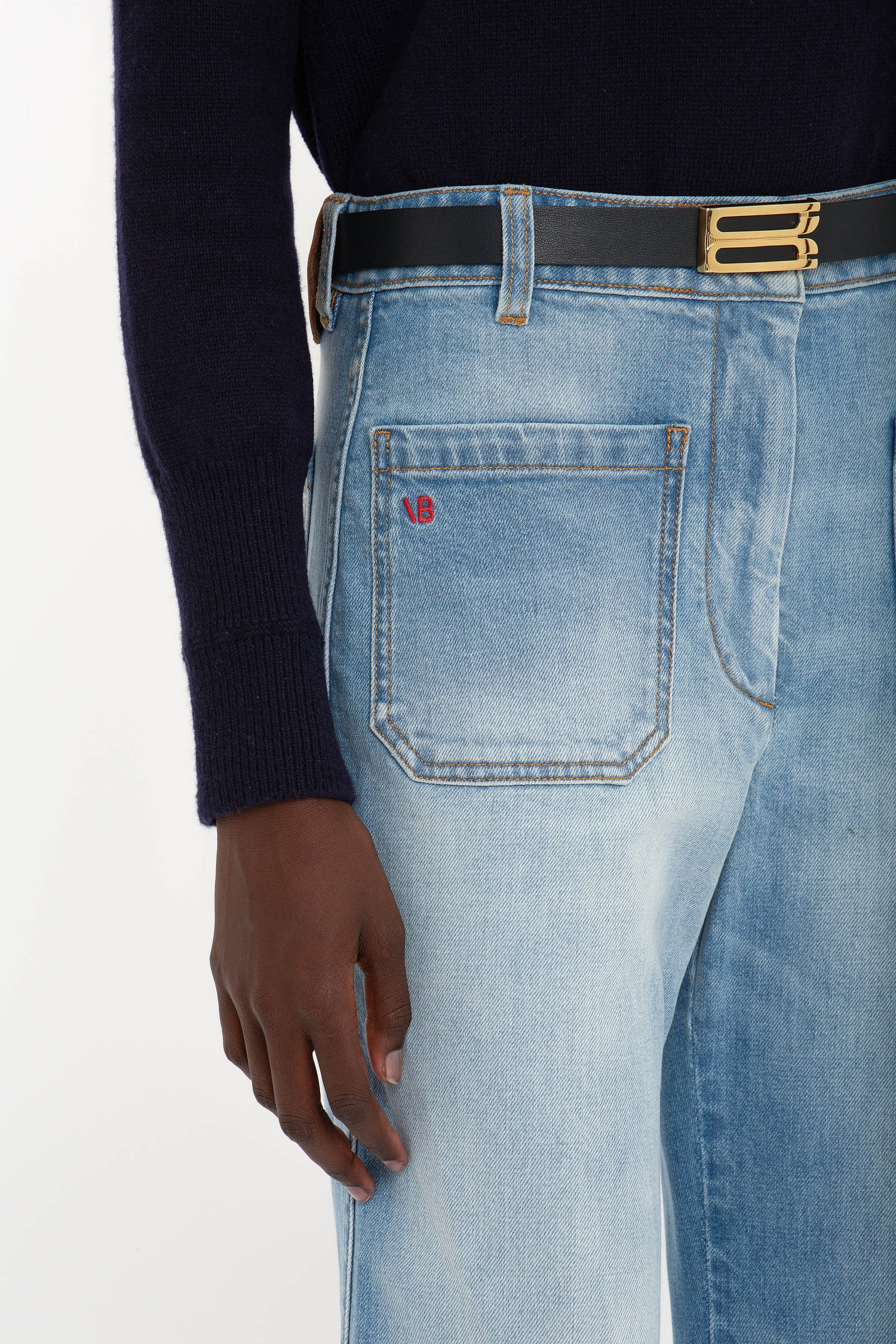 Person wearing Victoria Beckham Alina High Waisted Jean In Light Summer Wash with a red monogram detail on the back pocket, a black belt with a gold buckle, and a dark long-sleeve shirt. Only the lower torso and hand are visible.