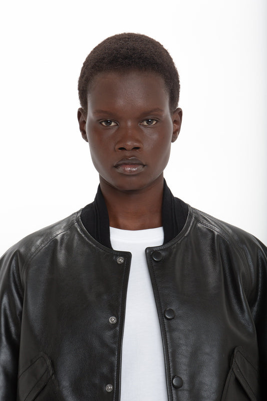 A person with a short haircut is wearing a Victoria Beckham Leather Varsity Jacket In Black over a white shirt, standing against a plain white background.