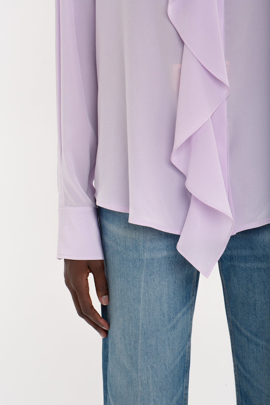 Person wears a petunia-hued Asymmetric Ruffle Blouse In Petunia by Victoria Beckham with blue jeans, pictured from the chest down against a plain background.