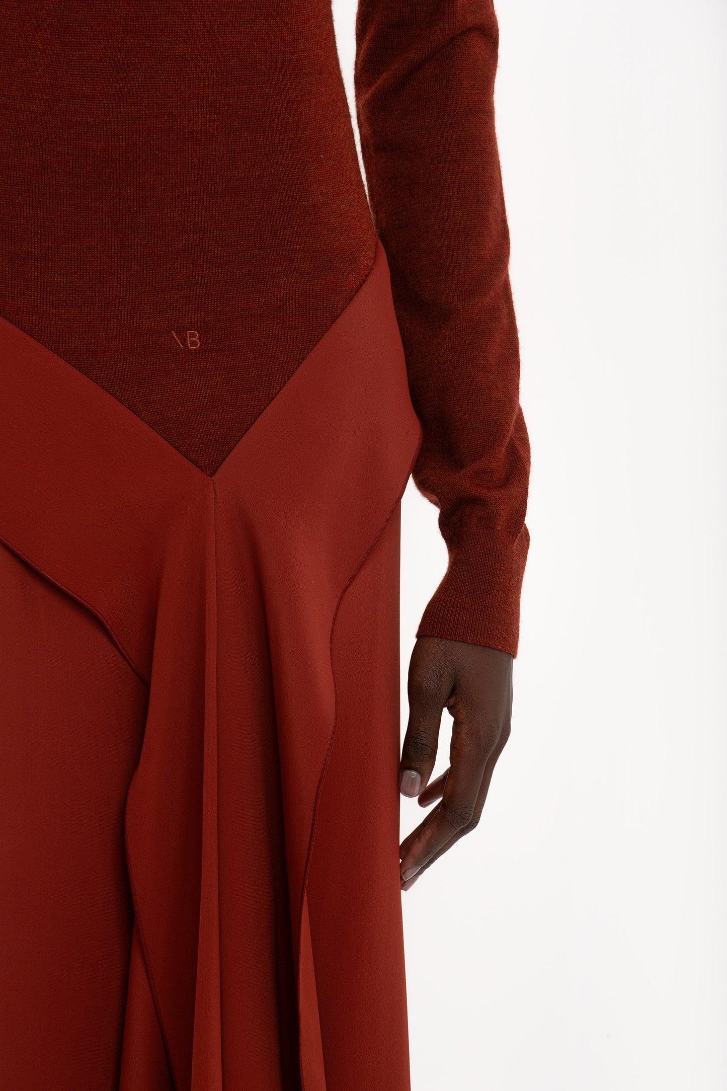 A close-up of a person wearing the High Neck Tie Detail Dress In Russet by Victoria Beckham. The person’s right arm is visible, and the fabric features pleats and an asymmetric hemline.