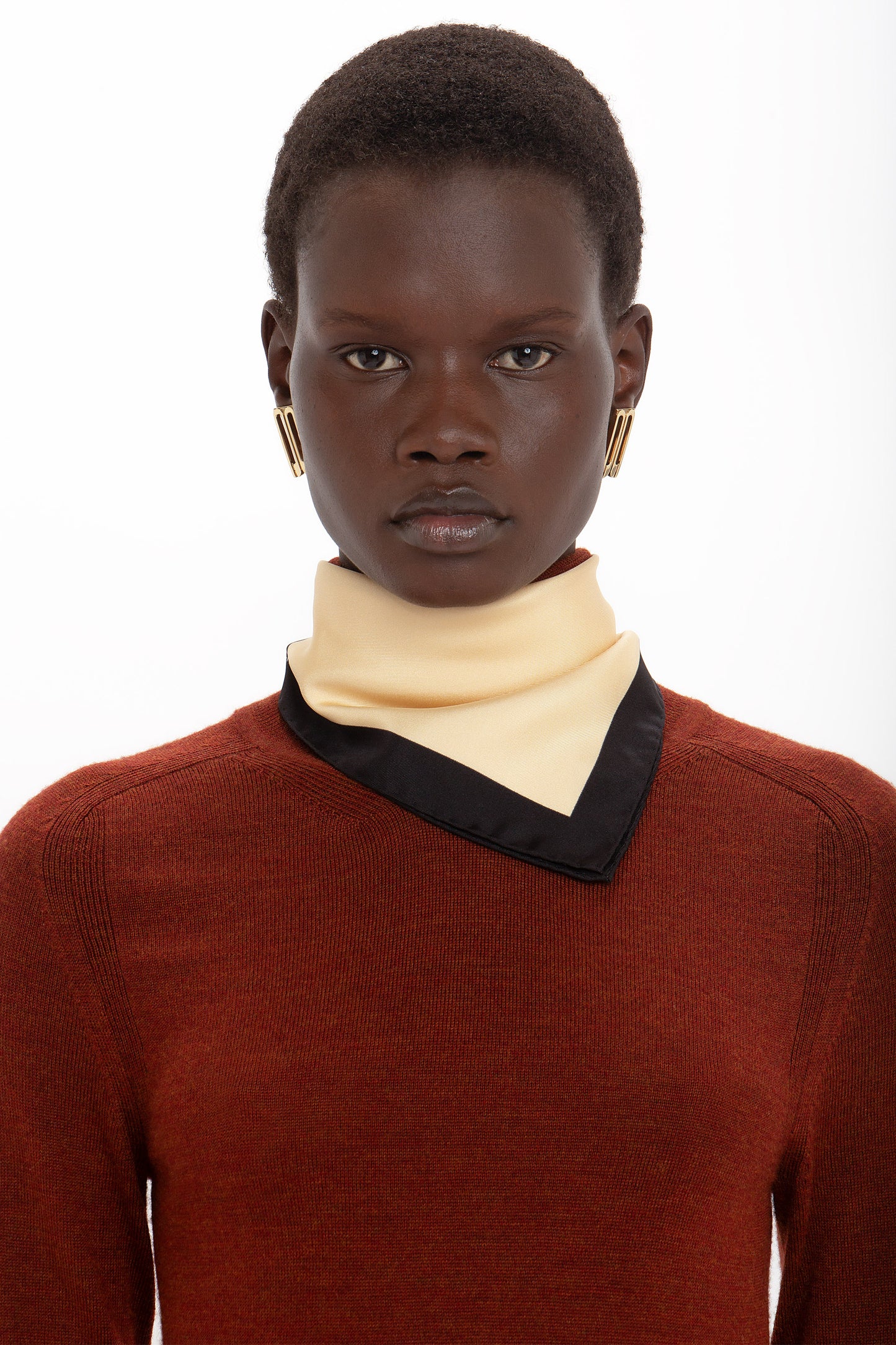 Person with short hair wearing a rust-colored sweater, a cream and black scarf, and gold geometric earrings, looking directly at the camera against a plain white background. The ensemble exudes sophisticated style akin to the Victoria Beckham High Neck Tie Detail Dress In Russet with merino wool bodices and asymmetric hemlines.
