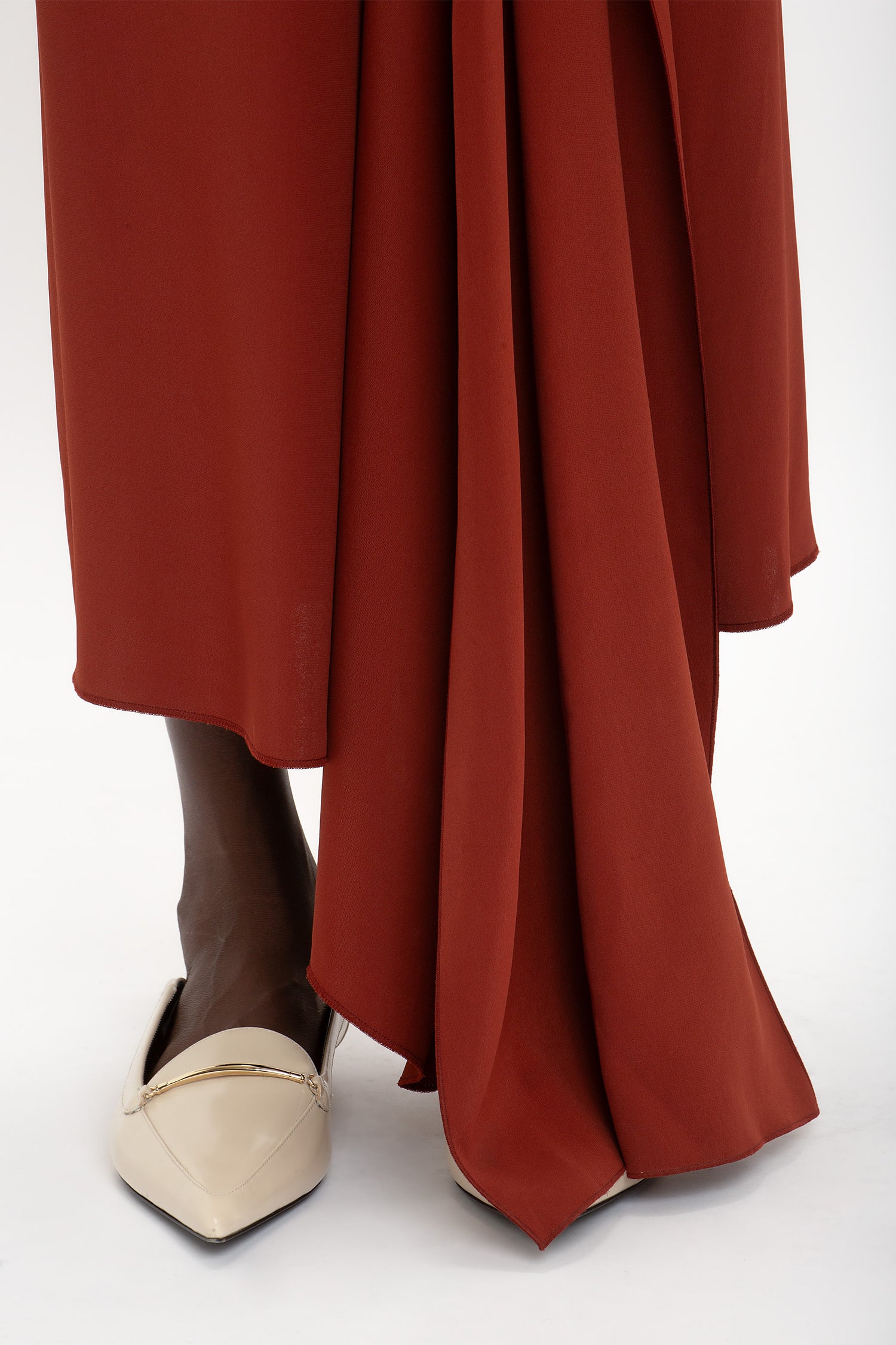 Close-up of a person wearing the Victoria Beckham High Neck Tie Detail Dress In Russet with an asymmetric hemline and beige pointed-toe flats.