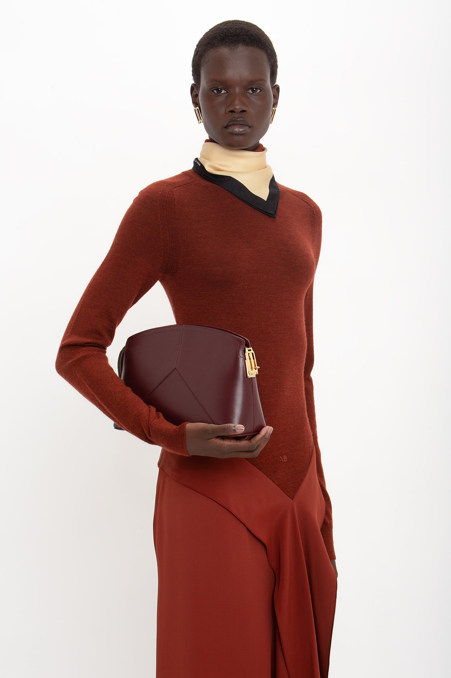 A person wearing a High Neck Tie Detail Dress In Russet by Victoria Beckham and carrying a maroon handbag stands against a white background.