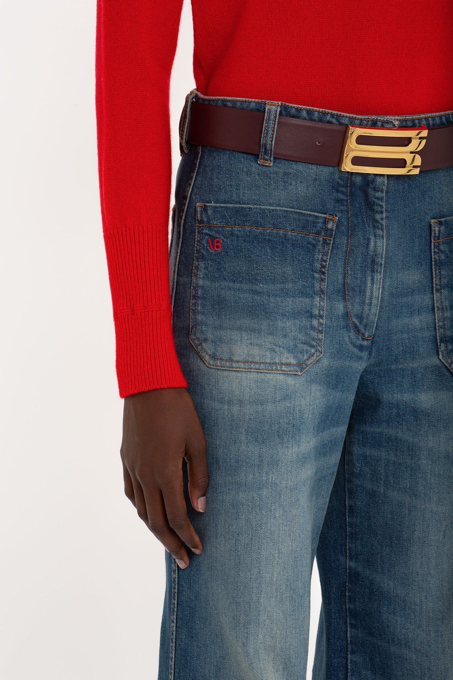 Person wearing a red long-sleeve shirt, Victoria Beckham's Alina High Waisted Jean In Heavy Vintage Indigo Wash with front pockets, and a brown belt with a gold buckle, standing against a white background, channeling Victoria Beckham’s retro appeal.