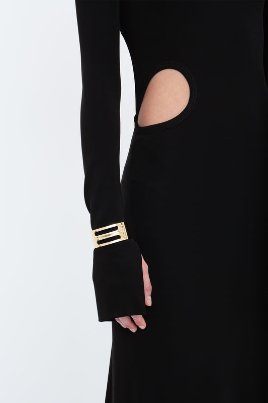Close-up of a person wearing the Victoria Beckham Cut-Out Jersey Floor-Length Dress In Black, accessorized with a gold bracelet on their wrist, reminiscent of the elegant evening gowns by Victoria Beckham.