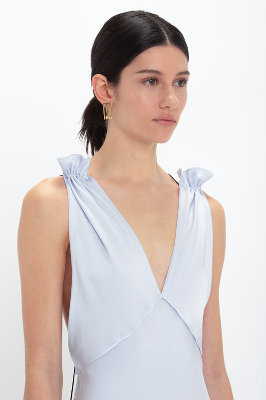 A woman with dark hair tied back, wearing an Exclusive Gathered Shoulder Cami Floor-Length Gown In Ice Blue by Victoria Beckham with a deep V-neckline and bow-like details on the shoulders, poses against a plain white background.
