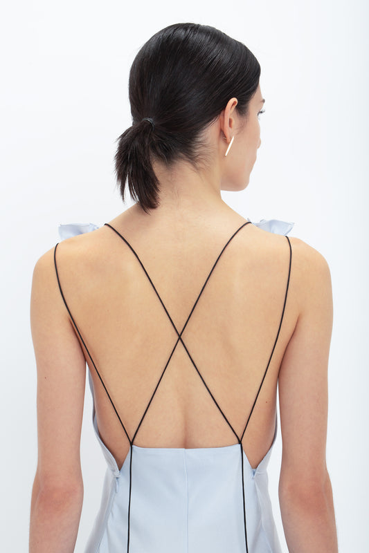 A woman with dark hair tied in a low ponytail is wearing the Victoria Beckham Exclusive Gathered Shoulder Cami Floor-Length Gown In Ice Blue, with thin black cross-straps. She is facing away from the camera.