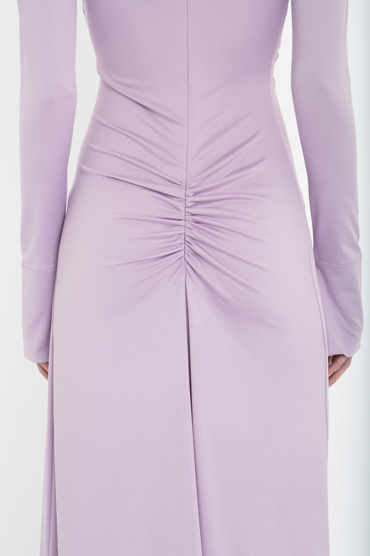 A close-up of the back of a person wearing a long-sleeved, fitted Victoria Beckham Ruched Detail Floor-Length Gown In Petunia in lavender stretch jersey with ruched detail at the waist.