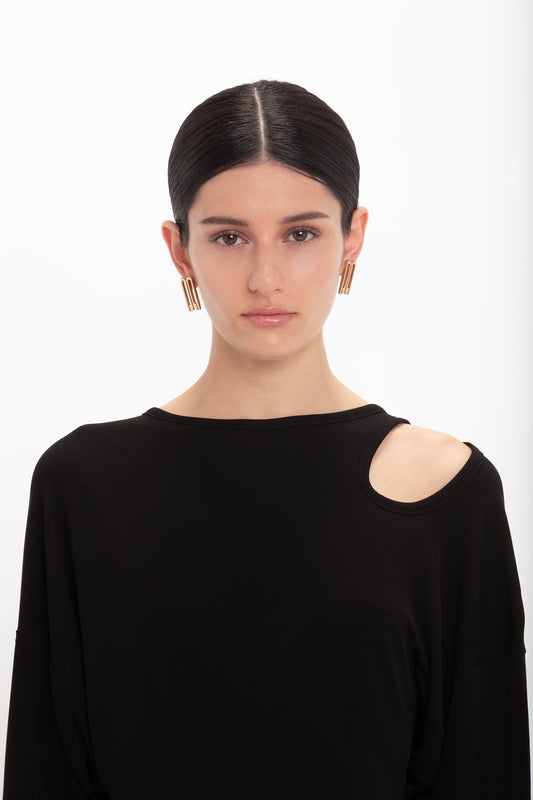 A person with dark hair, wearing gold teardrop earrings and a black Twist Detail Jersey Top In Black by Victoria Beckham with a cut-out shoulder, stands against a white background.