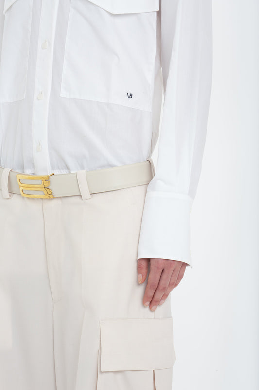 A person wearing a white long-sleeve shirt and military-inspired Relaxed Cargo Trouser In Bone by Victoria Beckham with a beige belt and gold buckle is shown from the torso down. The beige pants, made from 100% cotton, feature a stylish cargo pocket.