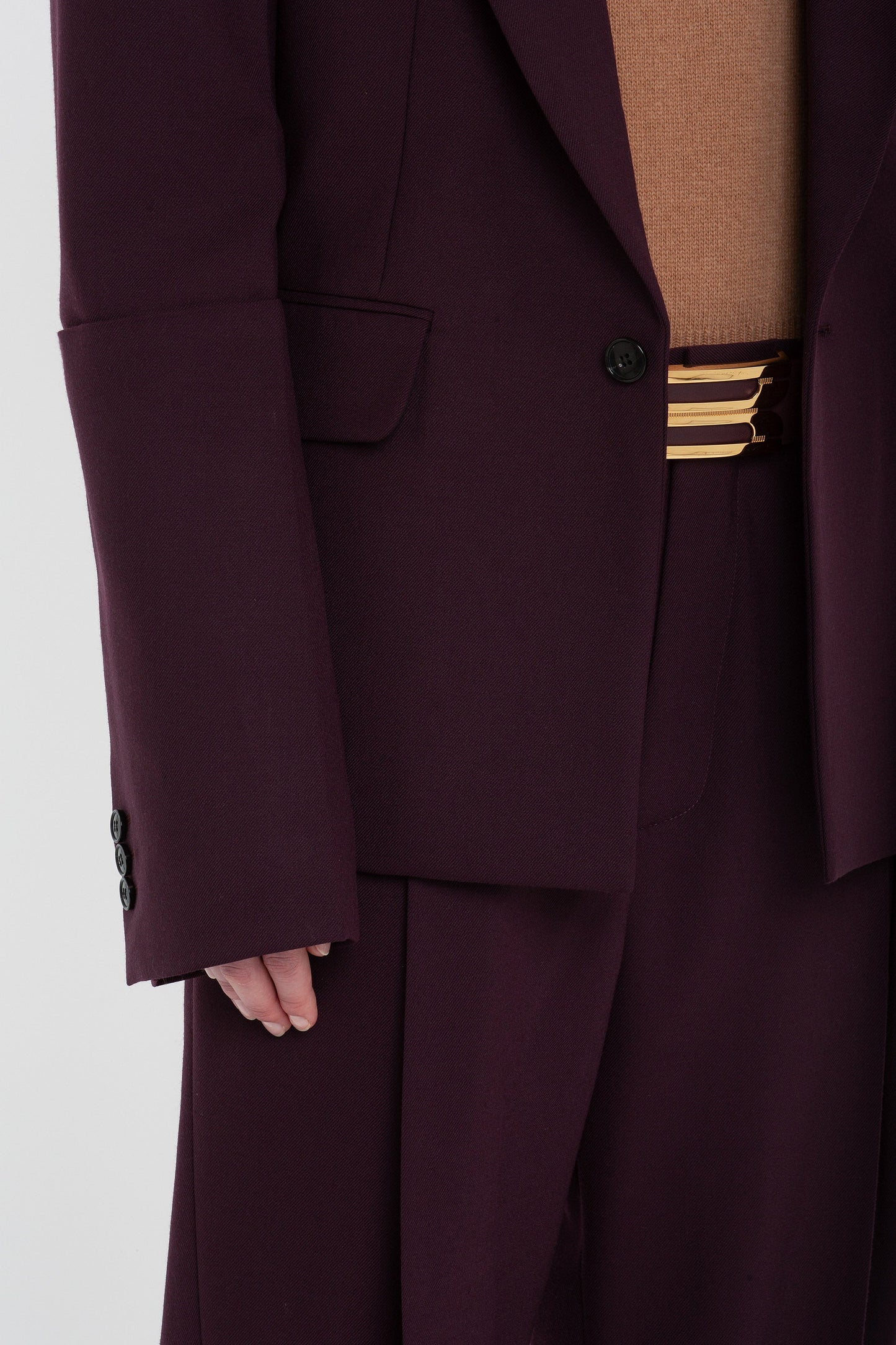 Close-up of a person wearing the Victoria Beckham Sleeve Detail Patch Pocket Jacket in Deep Mahogany with a brown sweater and matching pants, showcasing the black button detail on the sleeve and a striped belt.